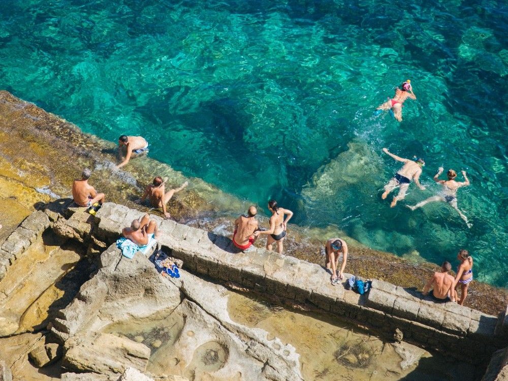 Una vecchia cava di tufo affacciata sul mare turchese, con persone che nuotano e prendono il sole sulla pietra