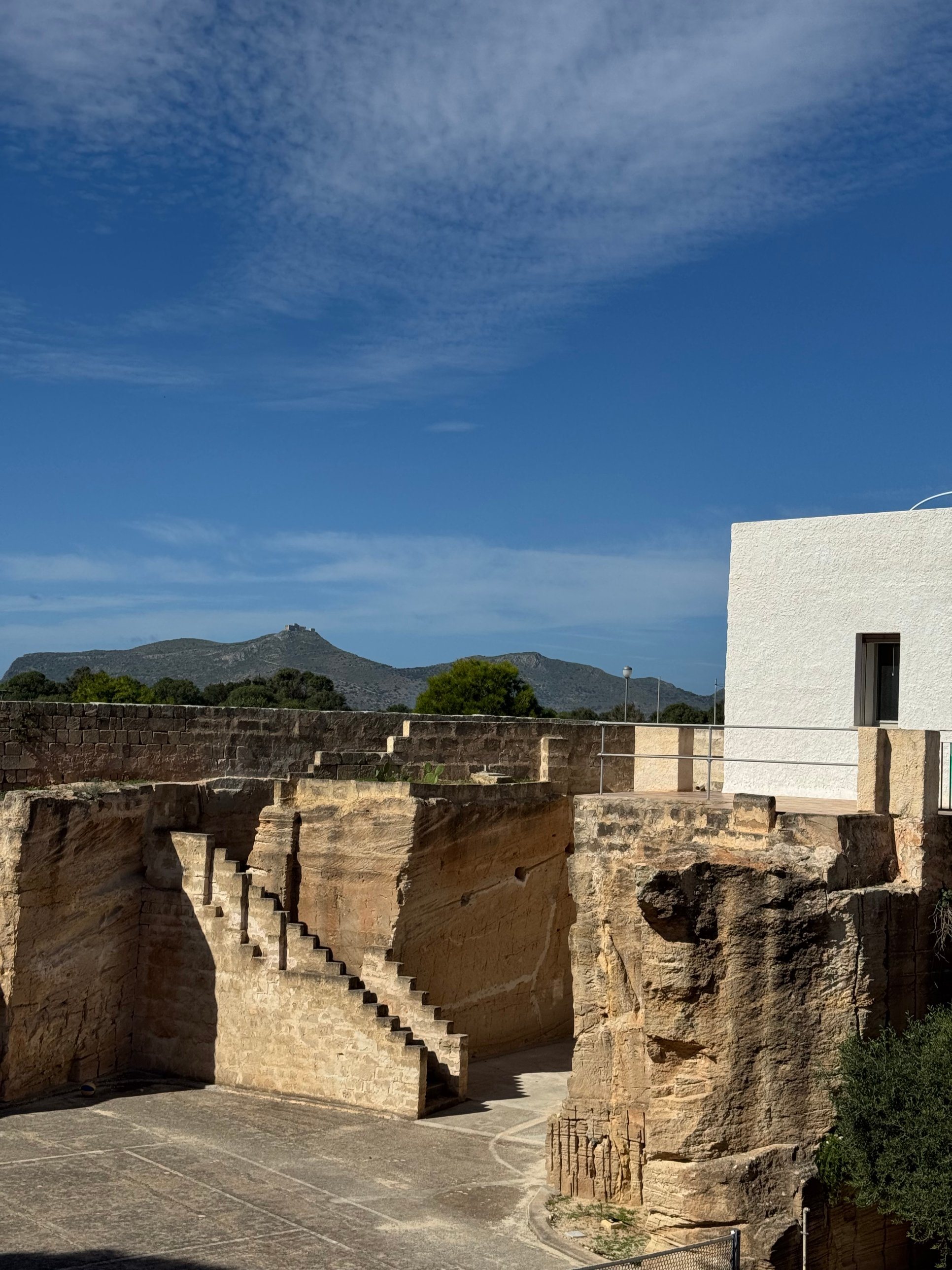 La villa bianca di MAE sopra la cava di tufo, con la scalinata scavata nella pietra e le colline sotto il cielo azzurro